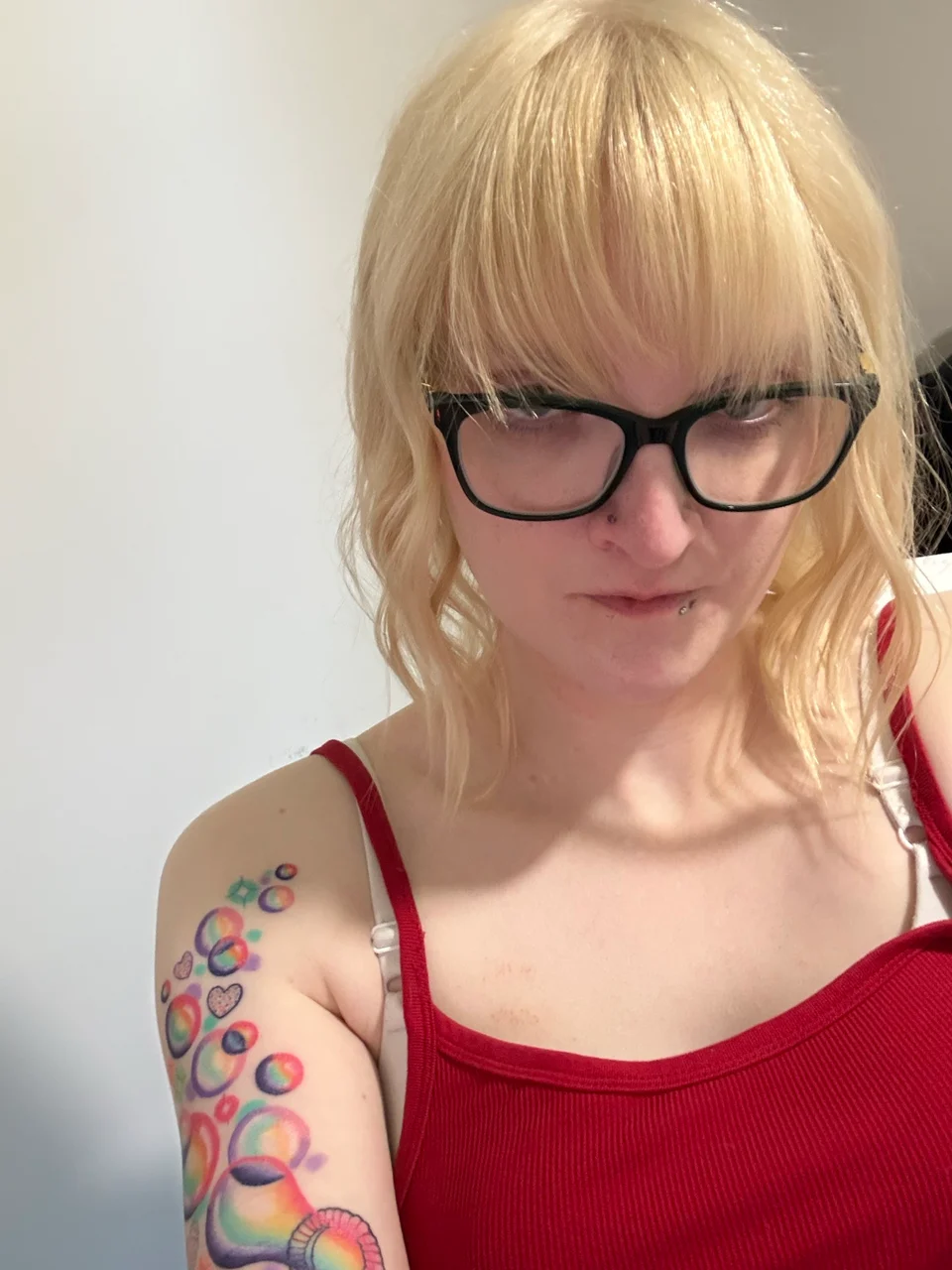 Alexis Everage with blonde hair, black glasses, a red top, and colorful bubble tattoos.